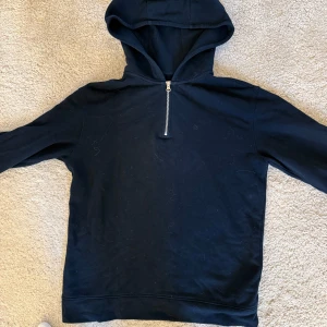 Mörkblå hoodie från East West - Säljer en stilren mörkblå hoodie från East West med dragkedja vid halsen. Perfekt för en avslappnad look. Den har en klassisk huva och långa ärmar. Storlek M. Mycket bra skick