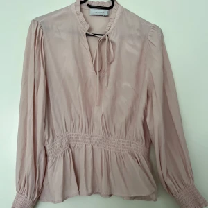 Blus, 365, rosa, S - Ljusrosa blus med långa ärmar och smockdetaljer i midjan och vid ärmslut, från 365 (MQ) i storlek S. Blusen har en liten krage och en v-formad öppning med knytband. Färgen är rosa. 100% viskos. I mycket fint skick.  Mått: Längd ca 60 cm Byst ca 48 cm Midja ca 34-55 cm (elastisk) Ärmlängd mätt från axeln ca 57 cm Axelmått ca 40 cm