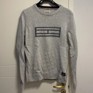 Grå stickad tröja från Hilfiger Denim - Säljer en stilren grå stickad tröja från Hilfiger Denim. Tröjan har ett diskret mönster på framsidan och ribbade muddar vid ärmslut och nederkant. Perfekt för kyligare dagar.