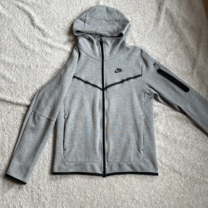 Grå nike tech hoodie, endast använd ett fåtal gånger - Säljer en grå hoodie från Nike med dragkedja och huva. Jackan har svarta detaljer och en ficka med dragkedja på.