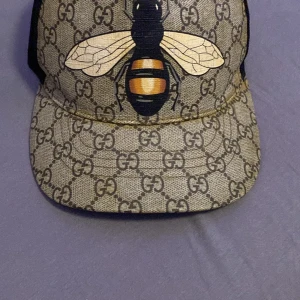 Beige keps med bi-motiv - Snygg beige keps från Gucci med ett stort bi-motiv på framsidan. Kepsen har det klassiska GG-mönstret och justerbart band med röd-gröna ränder. Perfekt för att ge din outfit en lyxig touch.