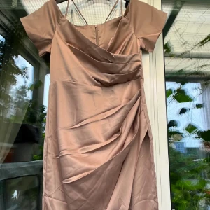 Champagnefärgad balklänning  - Elegant champagnefärgad klänning med korta ärmar och draperad detalj framtill. Klänningen har en v-ringad design och är tillverkad i ett glansigt material som ger en lyxig känsla.