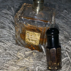 Khamrah Parfym från Lattafa - Elegant parfymflaska från Lattafa med namnet Khamrah. Flaskan har en fyrkantig form med ett intrikat mönster och en gyllene etikett. Parfymen har en djup bärnstensfärg. Den medföljande mindre flaskan har ett dekorativt mönster och en praktisk applikator.