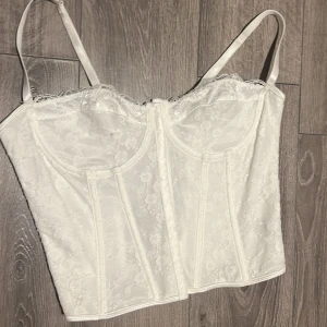 Vit spetskorsett med snörning - Säljer en superfin vit korsett med spetsdetaljer och justerbara axelband. Den har en snygg snörning i ryggen som ger en perfekt passform. Perfekt för fest eller speciella tillfällen!