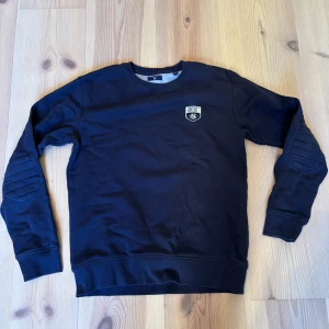 Svart sweatshirt från GANT - Säljer en stilren svart sweatshirt från GANT med ett broderat märke på bröstet. Tröjan har långa ärmar med ribbade detaljer vid armbågarna och ribbstickade muddar vid ärmslut och nederkant. Perfekt för en avslappnad look.