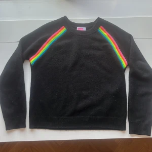 Svart tröja med regnbågsdetaljer från From Future - Säljer en svart tröja från From Future med färgglada regnbågsränder längs axlarna. Tröjan har långa ärmar och en rund halsringning. Frakt ingår inte i priset!