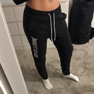 Svarta mjukisbyxor från Hollister - Säljer ett par svarta mjukisbyxor från Hollister med snörning i midjan och tryck längs benet. Perfekta för en avslappnad stil. Byxorna har en bekväm passform och är tillverkade i mjukt material.