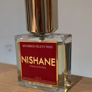 Hundred Silent Ways Extrait de Parfum från Nishane - Elegant och lyxig parfym från Nishane, 'Hundred Silent Ways'. Flaskan är fyrkantig med en röd etikett och gyllene detaljer. Doften är intensiv och sofistikerad, perfekt för den som vill sticka ut.