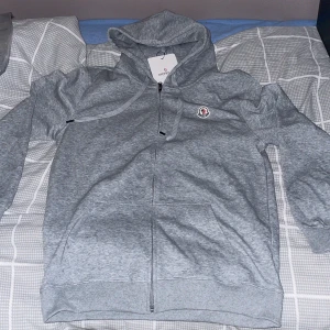 Grå hoodie från Moncler - Säljer en stilren grå hoodie  med dragkedja och huva. Tröjan har en broderad logotyp på bröstet och praktiska fickor framtill. Perfekt för en avslappnad look.