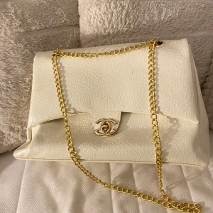Vit axelväska med guldkedja lyxig skolväska - Elegant vit axelväska med en stilren guldkedja och ett ikoniskt märke på framsidan. Väskan har en klassisk design som passar perfekt för olika tillfällen. Perekt för skolan!!