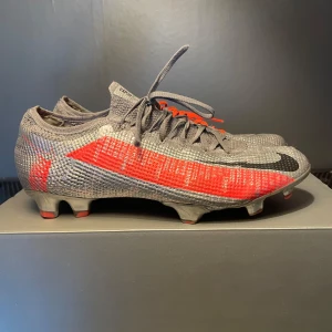 Grå och röda Nike Mercurial fotbollsskor - Snygga Nike Mercurial fotbollsskor i grått med röda och svarta detaljer. Skorna har en strömlinjeformad design med snörning och är perfekta för fotbollsplanen. De har en lätt och flexibel sula för optimal prestanda.