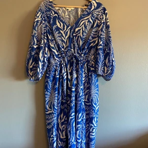 klänning - Säljer en blå och vit klänning från H&M med ett vackert bladmönster. Klänningen har en omlott-design och trekvartsärmar, vilket ger en elegant look. Perfekt för sommarens alla tillfällen!