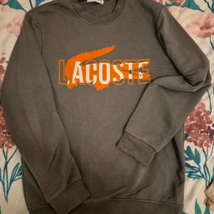 Grå sweatshirt från Lacoste - Säljer en grå sweatshirt från Lacoste med en stor orange och vit logotyp på framsidan. Tröjan har långa ärmar och rund halsringning. Perfekt för en avslappnad stil.