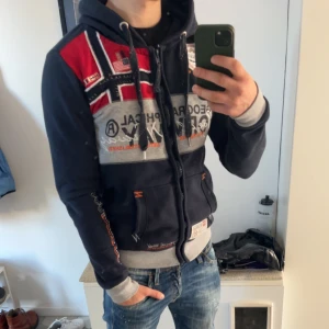 Mörkblå hoodie från Geographical Norway - Säljer en mörkblå hoodie från Geographical Norway med tryck och flaggdetaljer i rött och vitt. Tröjan har en justerbar huva med snören och ribbade muddar vid ärmslut och nederkant. Perfekt för en avslappnad stil. Ny pris på denna är 1299 kr 