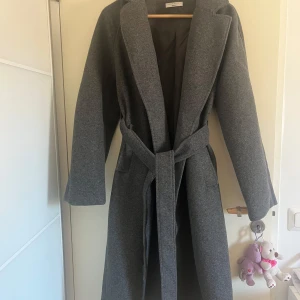 Grå kappa från Gina Tricot - Säljer en stilren grå kappa från Gina Tricot i storlek XS. Den har en klassisk design med knytbälte i midjan och långa ärmar. Perfekt för kyligare dagar.