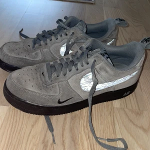 Grå Nike Air Force sneakers - Snygga grå Nike Air Force sneakers med svart sula och unikt mönstrad swoosh. Skorna har snörning och är tillverkade i mocka för en stilren look. Perfekta för en casual stil.
