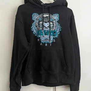 Svart hoodie från Kenzo med tigertryck - Säljer en autentisk svart hoodie från Kenzo med ett coolt tigertryck i blått och vitt på framsidan. Tröjan har en klassisk känguruficka och justerbar huva med dragsko. Perfekt för en avslappnad stil.