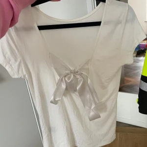 Vit kortärmad topp med knytband - Säljer en söt vit topp. Toppen har ett dekorativt knytband i satin som ger en fin detalj. Perfekt för en avslappnad stil.