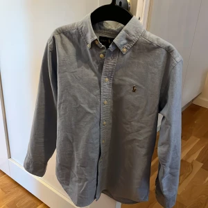 Ljus blå skjorta från Ralph Lauren - Snygg blå skjorta från Ralph Lauren med klassisk krage och knappar framtill. Skjortan har en broderad logotyp på bröstet och långa ärmar. Perfekt för en stilren look.