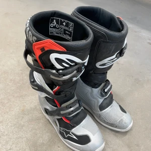 Svarta och grå motocrossstövlar från Alpinestars - Säljer ett par robusta motocrossstövlar från Alpinestars i svart och grått med röda detaljer. Stövlarna har flera spännen för säker passform och är designade för att ge optimalt skydd och komfort vid körning.