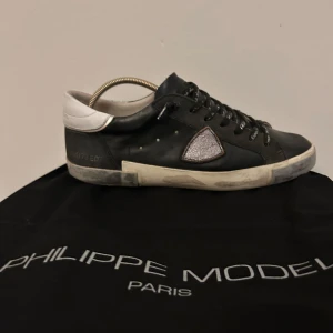 Svarta sneakers från Philippe Model - Snygga svarta sneakers från Philippe Model med vita detaljer och glittrig triangel på sidan. Skorna har snörning med text och en vit häl. Perfekta för en stilren look. Slitna på hälen men syns inte när man har på sig dem.