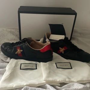 Svarta sneakers med bi-detalj från Gucci - Snygga svarta sneakers från Gucci med ikonisk bi-detalj på sidan. Skorna har röda och blå detaljer samt en kontrasterande häl i rött och blått. Perfekta för den stilmedvetna som vill ha något extra i garderoben. Med en spricka som man ser på bilderna