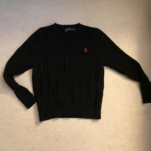 Svart stickad tröja från Ralph Lauren - Säljer en klassisk svart stickad tröja från Ralph Lauren med det ikoniska röda logotypbroderiet på bröstet. Tröjan har en ribbad struktur och långa ärmar, perfekt för kyligare dagar.