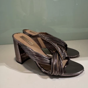 Silvriga klackskor från Saks Fifth Avenue - Snygga silvriga klackskor med öppen tå och korsade remmar. Perfekta för en kväll ute eller en speciell tillställning. Klackarna är breda och stabila, vilket ger både stil och komfort.