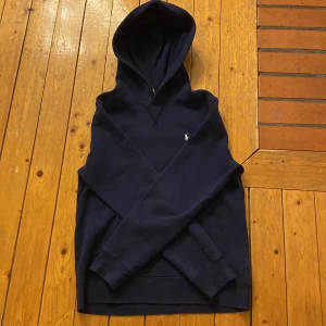 Mörkblå hoodie från Ralph Lauren - Säljer en stilren mörkblå hoodie från Ralph Lauren med en liten broderad logga på bröstet. Perfekt för en avslappnad look. Den har långa ärmar och en klassisk huva.