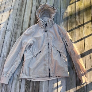 Beige vindjacka från C.P. Company - Snygg beige vindjacka från C.P. Company med huva och dragkedja. Jackan har långa ärmar och praktiska fickor med dragkedja. Perfekt för blåsiga dagar. Väldigt fin jacka med nypris på runt 5000 kr. Finns några fläckar som knappt syns. Försvinner i kemtvätt. Annars extremt bra skick