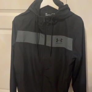 Svart vindjacka från Under Armour - Säljer en svart vindjacka från Under Armour med grått band över bröstet och dragkedja framtill. Jackan har en huva med dragsko och långa ärmar. Perfekt för träning eller vardagsbruk. Passar XS-S och är använd cirka 6 gånger