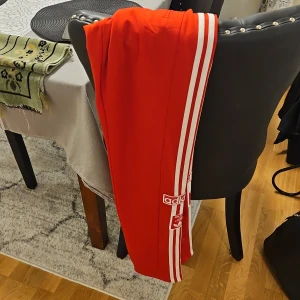 Röda träningsbyxor från Adidas - Snygga röda träningsbyxor från Adidas med klassiska vita ränder längs sidorna. Byxorna har knappar längs benen för justerbar stil och komfort. Perfekta för en sportig look.