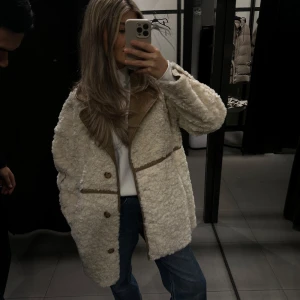 Beige teddyjacka med bruna detaljer - Säljer en mysig beige teddyjacka med bruna detaljer och knappar. Jackan har en normal passform och långa ärmar. Den har en stilren design med en skön krage och praktiska fickor framtill. Använd endast 1 gå så som ny, mer åt oversize så passar någon med S/M men då blir den bara lite mindre oversize