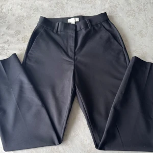 Svarta kostymbyxor (Högmidjade) från H&M - Eleganta svarta kostymbyxor från H&M med en klassisk passform. Byxorna har en stilren design med hällor för bälte och diskreta bakfickor. Perfekta för en stilren look.