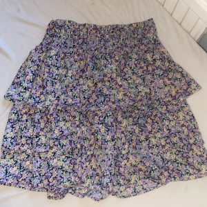 Blommig kjol från Vero Moda - Säljer en söt blommig kjol från Vero Moda i storlek M. Kjolen har ett färgglatt mönster i lila, gul och svart med en luftig passform. Perfekt för varma dagar!