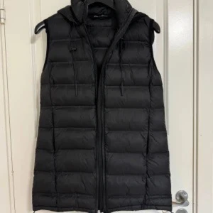 Zara svart väst  - Snygg svart dunväst med huva och dragkedja framtill. Perfekt för kyligare dagar och ger en sportig look. Västen har en quiltad design som ger extra värme och stil.