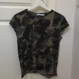 Camouflage t-shirt från NA-KD - Säljer en cool camouflage t-shirt från NA-KD med texten 'Feminist' broderad på bröstet. T-shirten är kortärmad och har en avslappnad passform. Perfekt för en casual look!