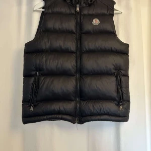 Svart dunväst från Moncler - Säljer en snygg svart dunväst från Moncler med dragkedja och logotyp på bröstet. Västen har en quiltad design och praktiska fickor med dragkedjor. Perfekt för kyliga dagar.