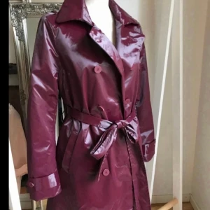 Vinröd trenchcoat från K-Way - Snygg vinröd trenchcoat från K-Way med knappar och bälte i midjan. Jackan har en glansig finish och långa ärmar, perfekt för höst och vår. Den har en klassisk krage och är tillverkad i ett syntetmaterial.OBS! Jackan gnisslar något vid rörelse pga materialet. Men otrolig snygg och skön jacka!  Perfekt för kallt o blött väder då den är fodrad! Använt 2 ggr max.I fint skick!