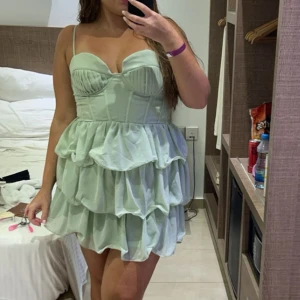 Ljusgrön klänning med volanger - Säljer en söt ljusgrön klänning med smala axelband och volanger. Klänningen har en hjärtformad byst och en kort, lekfull kjol med flera lager volanger. Perfekt för sommarens festligheter!
