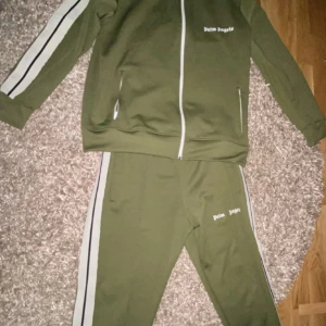 Olivgrön dress från Palm Angels helt ny använd 2 gånger äkta  - Snyggt olivgrönt dress från Palm Angels med vita detaljer. Jackan har dragkedja och vita ränder längs ärmarna. Byxorna har matchande ränder längs sidorna. Perfekt med bra kvalité och äkta kan även sänka pris. För mer information kontakta mig.