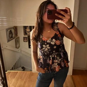 Blommig linnetopp - Blommig linnetopp med breda axelband och volangdetalj i nederkant. Toppen har ett färgglatt mönster med inslag av orange, lila och vitt på en mörk bas. Blusen är från Gina tricot i storlek s