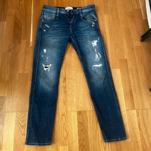 Blå jeans från Replay - Snygga blå jeans från Replay med slitna detaljer och en klassisk femficksdesign. De har en knappgylf och är perfekta för en avslappnad stil. Märkeslappen syns tydligt på baksidan. Storleken är 30/30 Anbass. 