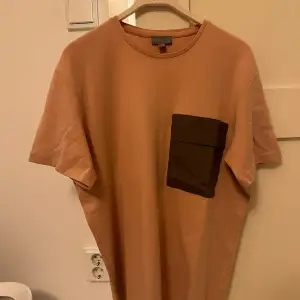 Säljer en stilren rosa t-shirt med en kontrasterande mörk ficka på bröstet. Perfekt för en avslappnad look. T-shirten har korta ärmar och en rund halsringning.