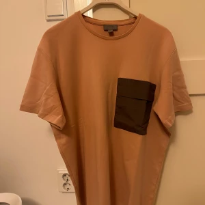 COS Rosa pink t-shirt med ficka - Säljer en stilren rosa t-shirt med en kontrasterande mörk ficka på bröstet. Perfekt för en avslappnad look. T-shirten har korta ärmar och en rund halsringning.