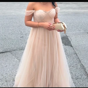 Champagnefärgad axelbandslös långklänning - Elegant champagnefärgad långklänning med offshoulder-design och v-ringad urringning. Klänningen har ett vackert fall och är perfekt för festliga tillfällen. Den är tillverkad i ett lätt och luftigt material som ger en drömlik känsla.