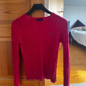 Rosa stickad tröja från Ralph Lauren - Säljer en snygg rosa stickad tröja från Ralph Lauren med kabelstickat mönster. Tröjan har långa ärmar och en liten grön logga på bröstet. Perfekt för kyligare dagar!
