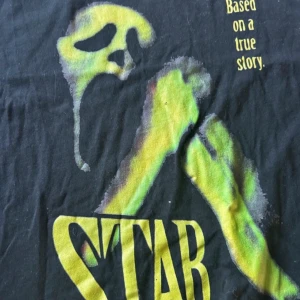 Svart t-shirt med tryck Stab Scream - Cool svart t-shirt med ett stort tryck av en skrikande mask i grönt och gult. Texten 'STAB' och 'Based on a true story' ger en mystisk känsla. Perfekt för filmälskare eller de som gillar unika tryck.