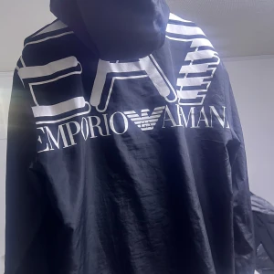 Svart vindjacka från Emporio Armani EA7 - Säljer en svart vindjacka från Emporio Armani EA7 med stor logga på ryggen och mindre logga på bröstet. Jackan har huva och dragsko nertill för justerbar passform. Perfekt för dig som gillar stilrena och sportiga plagg. Dragsko sönder på höger sida.. samt att dragkedja för att få in luvan har hängt sig