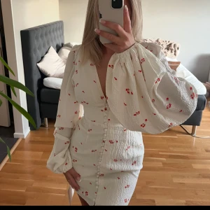 Adoore Vit kortklänning  - Supersöt vit kortklänning med små röda blommor och markerad midja. Klänningen har djup v-ringning, knappar framtill och långa puffärmar med resår vid handlederna. Perfekt för sommarens alla tillfällen!använd 1 tillfälle